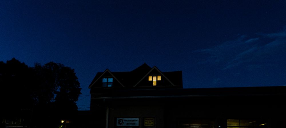 A nighttime scene featuring a dark blue sky above a small building with a triangular roof. One upper window glows with warm yellow light, while the rest of the structure remains dim. A sign reading “Amy’s Automotive” is visible below, along with business hours and a phone number. Trees frame the left side, and the quiet atmosphere gives you closure and solitude.
