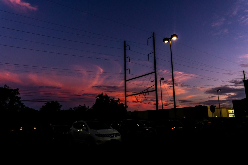 A twilight parking lot bathed in soft sunset hues. The sky shifts from purple to pink to orange, layered like quiet data. Streetlights flicker on against silhouetted power poles and stationary cars. The atmosphere feels held~neither day nor night, just transition behaving.
