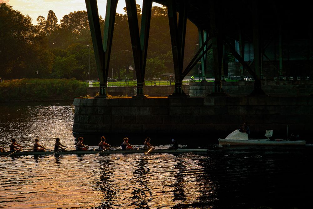 Orange hues flood the water’s surface, reflected from a concentrated morning sky. A narrow boat moves through the light—each rower precise, not decorative. The bridge above holds structural weight, not framing. Trees and grass line the far bank, unlit but intact. The orange operates as active field—held in water, sky, and motion.
