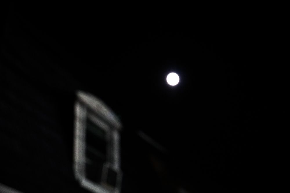 A night scene featuring a luminous full moon against a dark sky. In the foreground, a portion of a building with a softly blurred window adds depth and contrast, emphasizing the moon’s brilliance.
