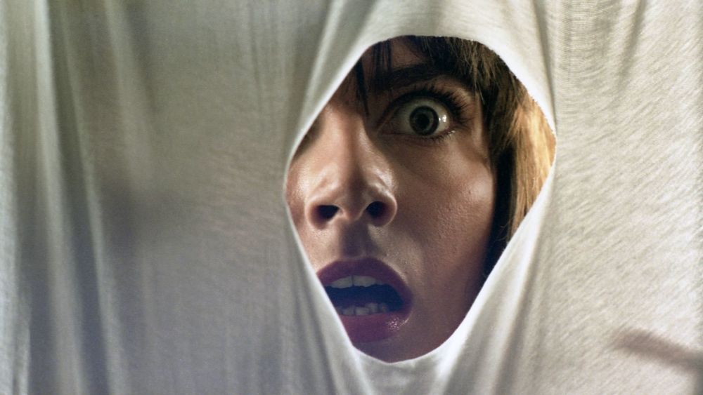 A woman stares in shock, eyes wide and mouth open, through a white cloth torn open to reveal her face