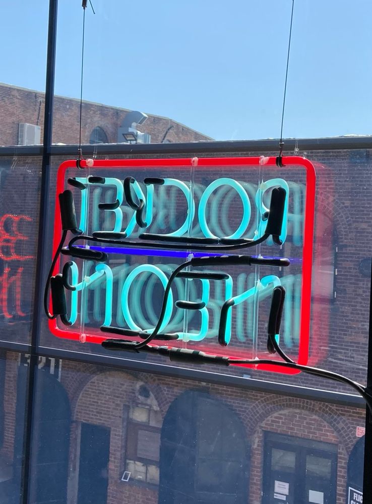 Neon sign hanging in window that reads, "ROCKET NEON" in turquoise lettering with purple and red borders. Bright blue sky seen in the background.