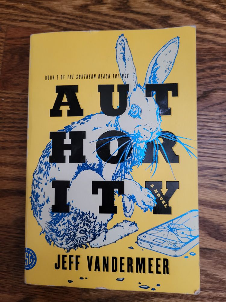 Photo of the cover of authority by Jeff Vandermeer. The cover is yellow and has a bunny with a cracked cell phone on it. 