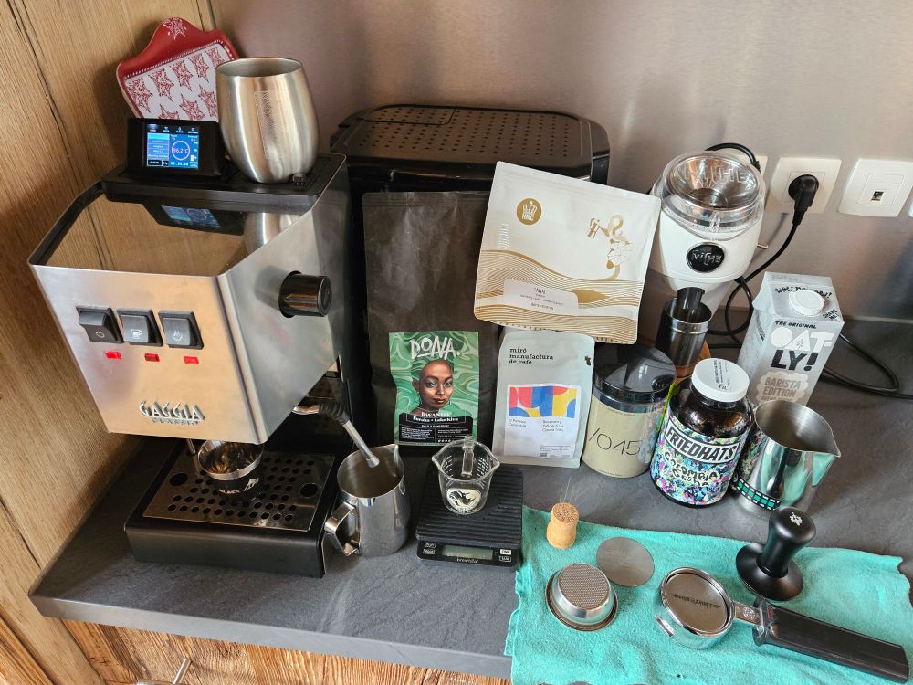 A nice espresso setup, with a Gaggiuino (modded Gaggia classic) on left, followed by a bunch of espresso tools and a bunch of bags of coffee beans, with a white Niche Zero grinder in the back and a naked portafilter on the front. 