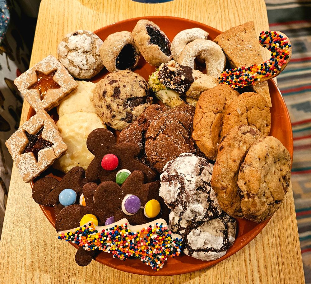 A plate of homemade cookies. Some are covered in sprinkles or sugar, there are jammy cookies, gingerbread, shortbread, sugar cookies. Many different kinds, colours and shapes.