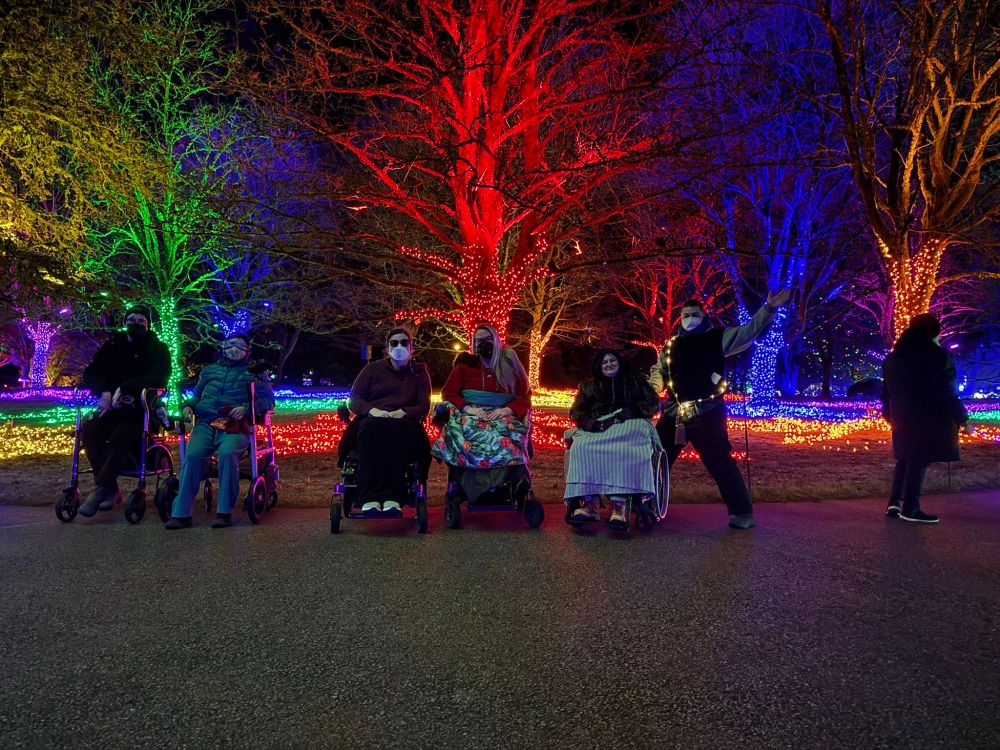 Six masked humans sitting or standing with their mobility aids. They're all masked, and standing in a row in front of a stand of brightly coloured trees. It's nighttime and the trees are lit up in the colours of the rainbow. We called these the "queer trees" so obviously we had to get a picture in front of them!