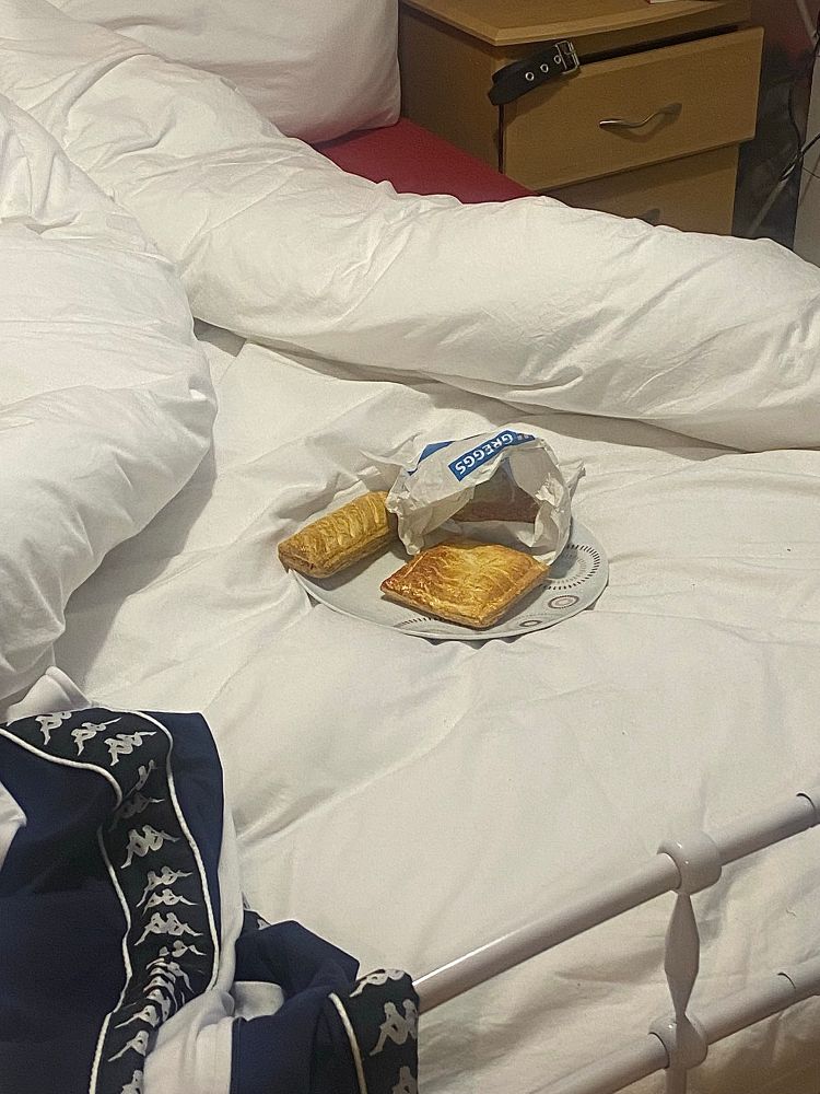 A bed with simple white bedding. A Kappa tracksuit thrown on the side. In the middle, an unpacked Greggs bag with two (?) pastries on a plasticky plate. 