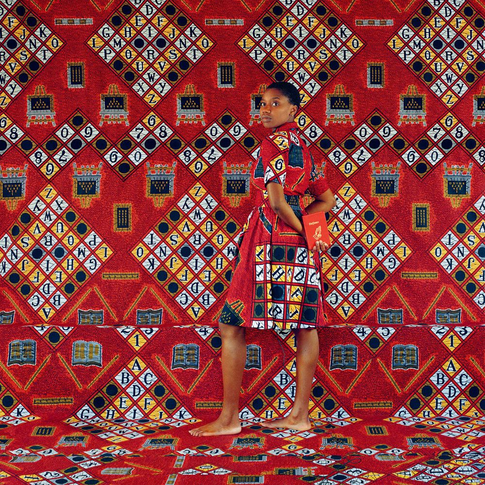 Color photo of a young Black woman standing against a bright red backdrop with a diamond pattern of black letters within yellow and white diamonds. She is wearing a dress that matches the backdrop so she partially blends in, and holds a red book behind her back while looking back at the viewer.