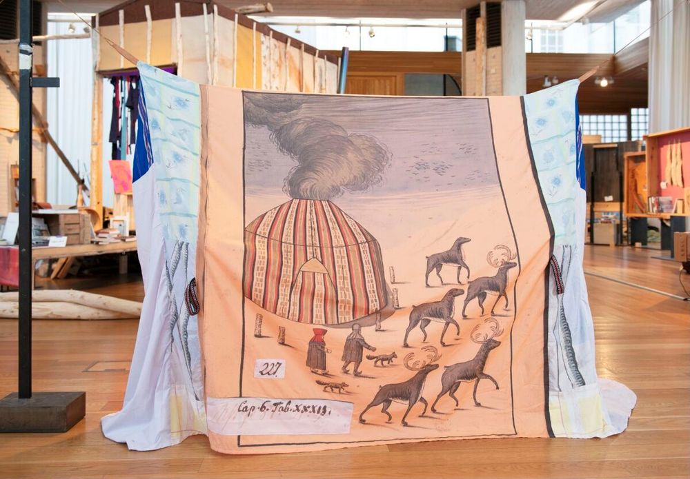 Sculptural tent structure made out of a blue textile suspended by thin ropes, decorated with an illustration of a striped hut with smoke emitting from its ceiling, with a group of stags and two human figures in front.