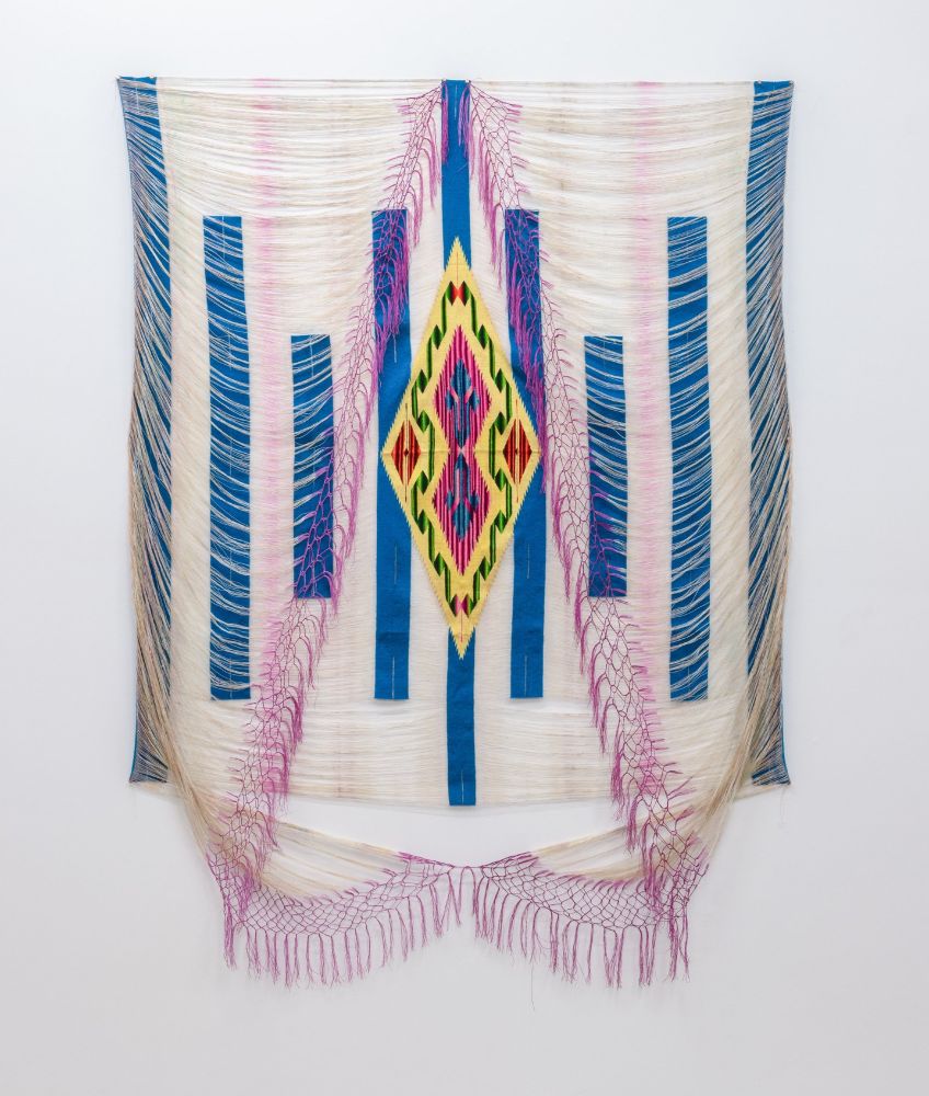 Hanging textile with blue vertical stripes and loose white threads draping in front with pink edges. A slender diamond with a yellow, pink, and green pattern sits in the center.