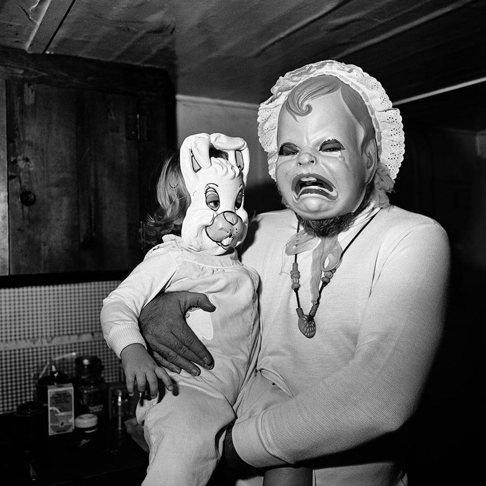 Black and white photo of a figure wearing a crying baby mask, holding a toddler wearing a bunny mask, standing in a room with a low ceiling and table with food jars.