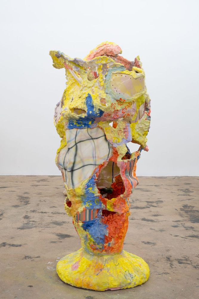 Standing abstract sculpture with a rounded yellow base and textured cylindrical shape moving upward with undulating curves and section sof cutouts and sections of translucent plastic.
