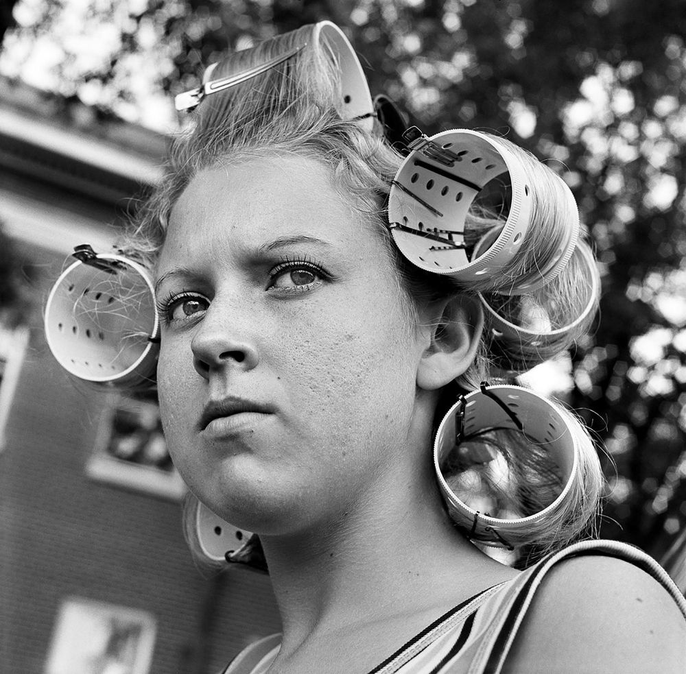 Black and white photo of a white teen girl with huge rollers in her blonde hair, shot in close-up outside a house with a large tree behind her.