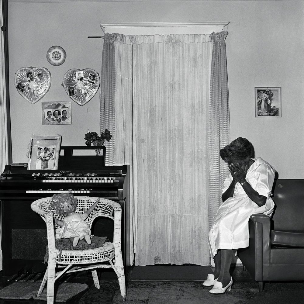 Black and white photo of a Black woman sitting on the edge of a couch, her head in her hands, in a room with a large window curtain and photos on the walls. A white wicker chair holding a blonde doll sits across from her with a small piano against the wall.