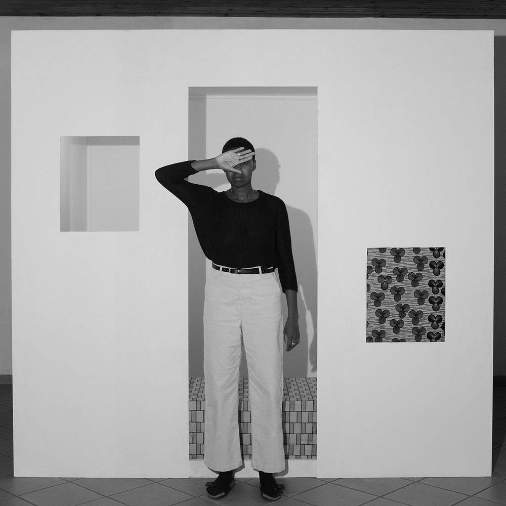 Black and white photo of a young Black woman wearing a dark shirt and light pants, standing in the center with her right hand held up against her face as if blocking bright light. Behind her is a white wall with cut-out rectangles.