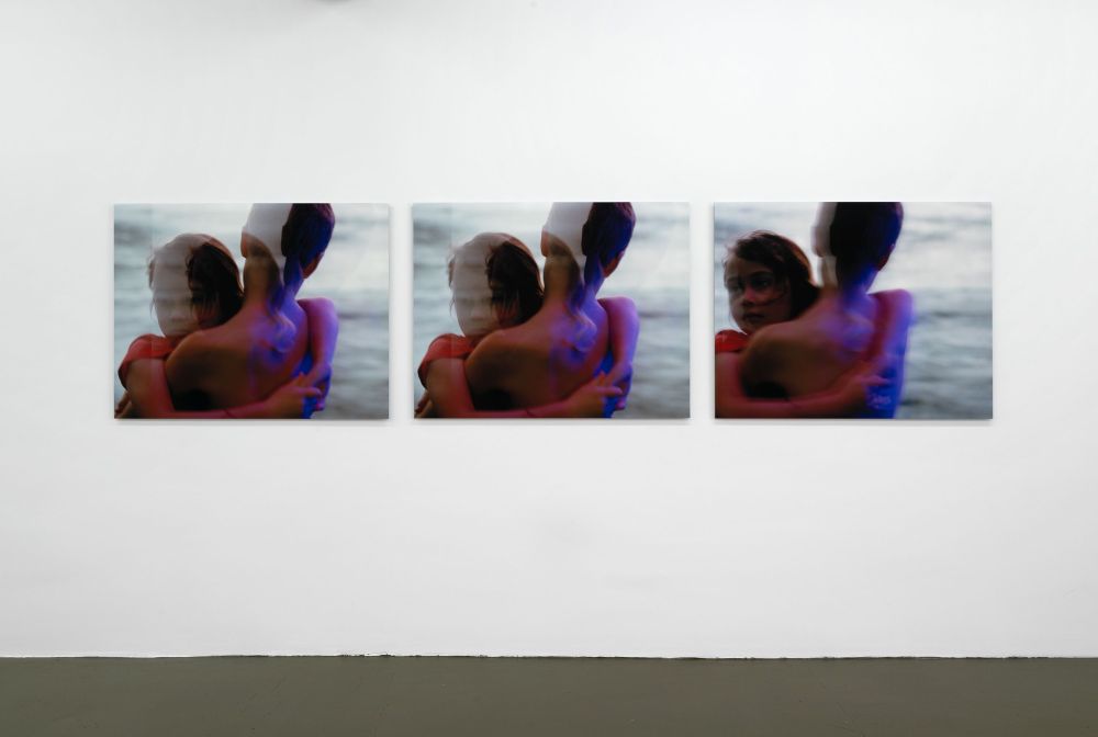 A gallery wall with three photos in a horizontal line, each depicting a similar composition of two young figures hugging with the ocean behind them, with overlaid elements slightly blurring the image and elements of red/purple/blue light.
