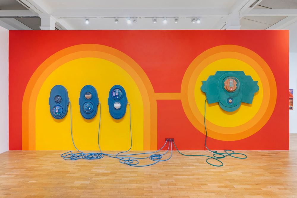 Gallery installation of a large wall painted red-orange with yellow and blue circular shapes. Blue and teal abstract forms hang on the wall, with long blue cords falling to the floor and plugging into the wall.