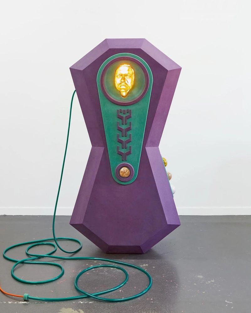 Abstract standing sculpture in an hourglass shape, painted deep purple with green and gold details, including a gold image of a man's face.