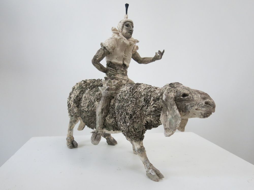 Clay sculpture of a human figure with a raised hand as if in conversation, sitting astride a large goat.