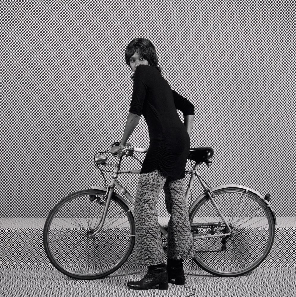 Black and white photo of a young Black woman looking over her shoulder at the viewer, standing and holding a bicycle, against a diamond patterned backdrop.