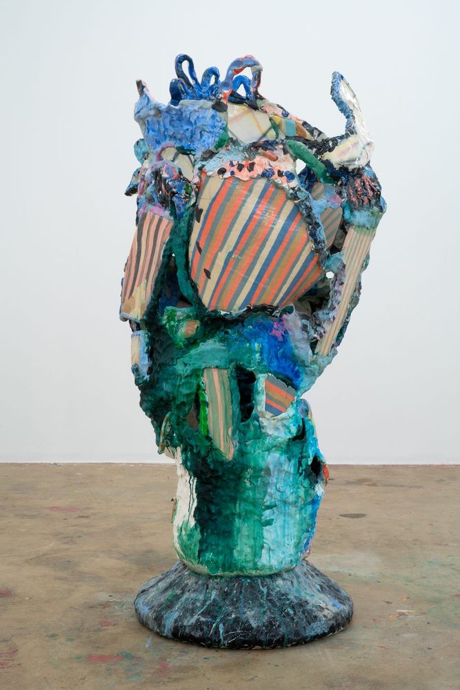 Standing abstract sculpture with a rounded base, a cylindrical green bottom, and a mass of shapes and colorful objects jumbled together along the upper half.
