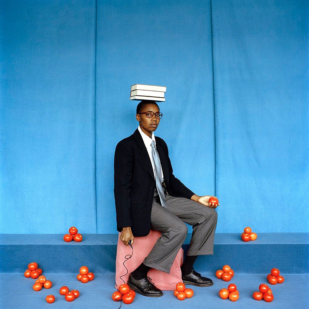 Color photo of a Black woman dressed in a men's suit, sitting on a pink block against a light blue background. She balances books on her head and holds a tomato, with several small piles of tomatoes surrounding her feet.