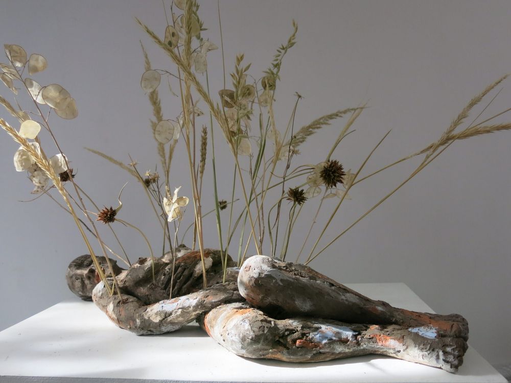 Clay sculpture of a woman lying on a white platform with her legs curled up and her hands on her torso. Dried flowers and tall grasses spring up from her body.