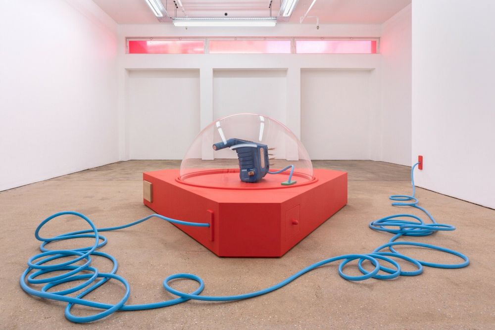 Sculpture installation of a large red-orange platform holding a blue abstract machine sitting within a clear plastic dome, powered by a long thick blue electric cord twisting through the gallery.