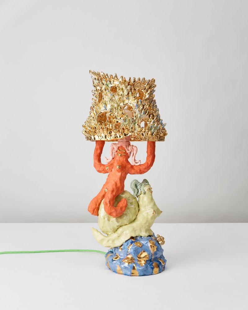 Lamp sculpture of a green snail-woman hybrid sitting atop a blue and gold blob, with an orange female figure on top of the snail's shell holding a textured gold lampshade above her head.
