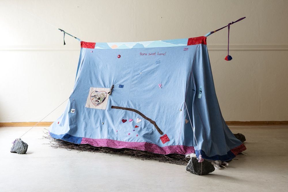 Sculptural tent structure made out of a light blue textile with embroidery, held up by thin rope lines, with thin twigs along its floor.