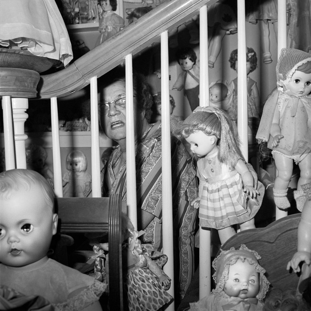 Black and white photo of a woman sitting behind a staircase banister with her eyes closed and mouth open, surrounded by dozens of white dolls.