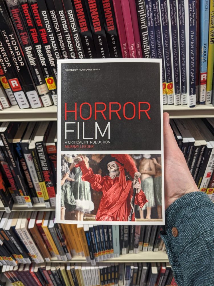 Someone holding a copy of the book 'Horror Film: a critical introduction' by Murray Leeder, in front of two shelves of various horror-related books inside Northumbria University City Campus Library.