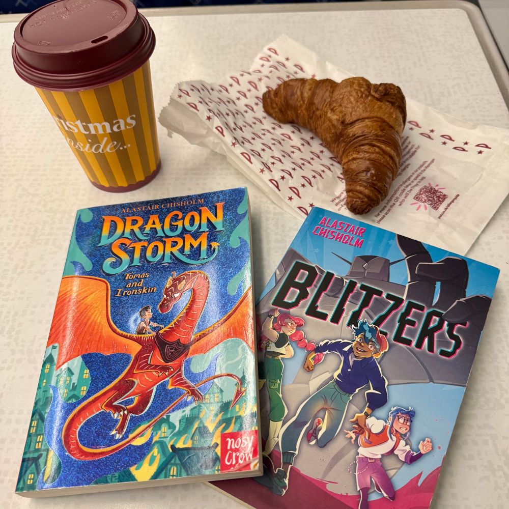 A train table with a coffee (top-left), a croissant (top-right), a copy of the book DRAGON STORM TOMAS AND IRONSKIN by Alastair Chisholm (bottom-left) and a copy of the book BLITZERS by Alastair Chisholm (bottom-right).