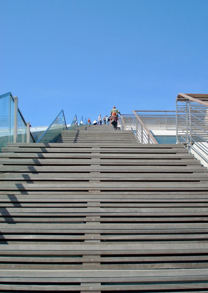 Eine breite hölzerne Treppe mit transparenten Geländern führt nach oben in Richtung des blauen Himmels. Auf den oberen Stufen stehen vereinzelt Menschen.