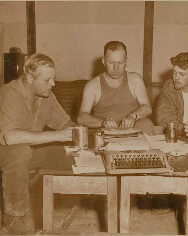 Photograph of Randolph Churchill and other war correspondents sitting round a table with typewriters, notes and drinks