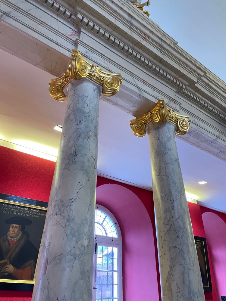 Graue Marmorsäulen mit goldener Verzierung vor einem großen Fenster und tief roter Wand mit Gemälden.