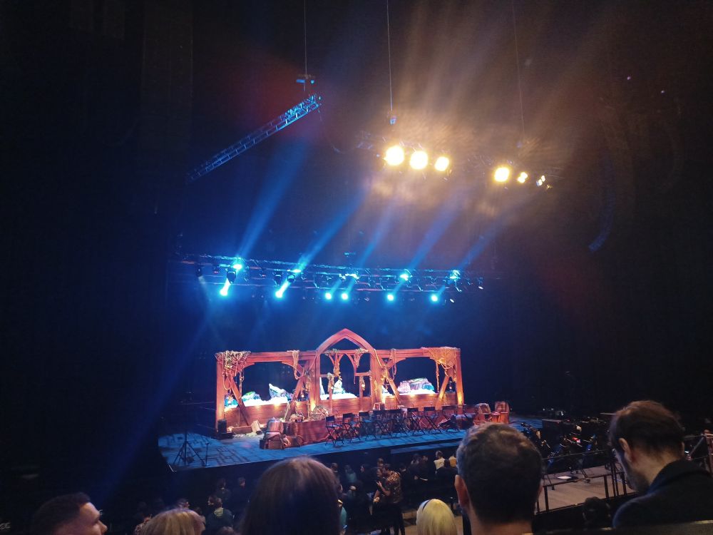 a distant picture of the critical role sydney live show stage. it is before the q&a session, so right chairs are set up in front of the orange setpiece. blue stage lights are overhead, and yellow theatre lights illuminate audience members in the bottom part of the image.