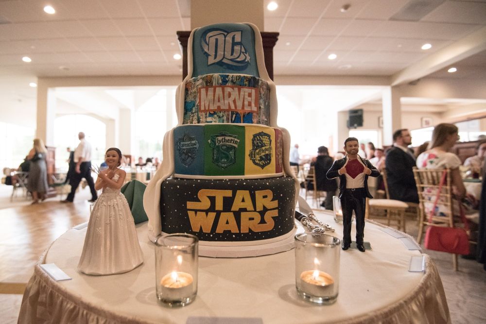 A four-tiered wedding cake with frosting that pulls back like a curtain. Each tier features a different logo: the top has a DC logo, the second has a Marvel logo against panels of Mister Fantastic and Invisible Woman's wedding, the four tier has the four Hogwarts houses, and the last tier has the Star Wars logo against a field of stars. The topper shows the bride with her arms crossed and Wolverine claws, while the groom opens his shirt to reveal a Daredevil logo.