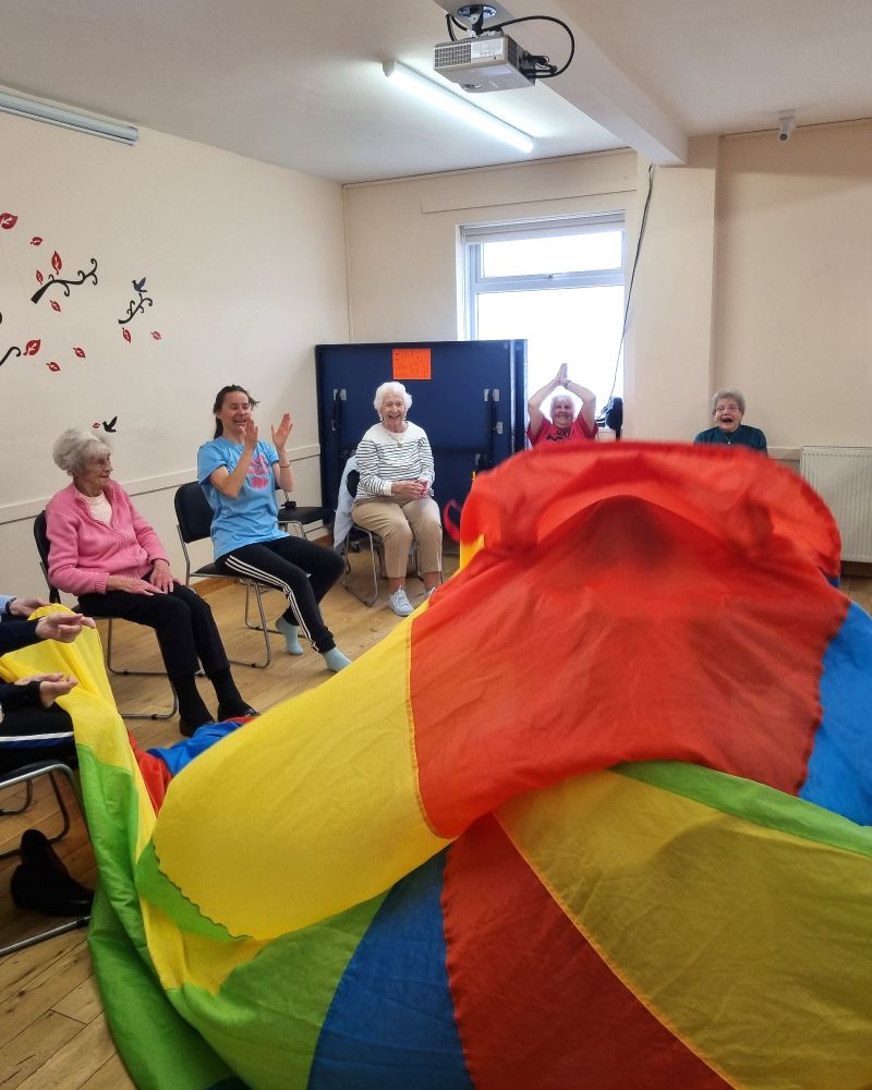 Photo of a parachute falling whilst Dove Centre service users smile, wave and clap during their gentle exercise class. 