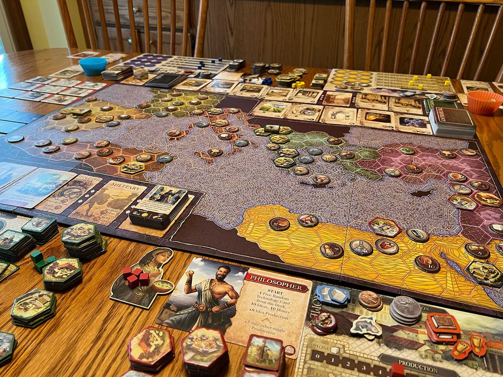Photo of the game Mosaic, showcasing a map with several tokens on landmasses that are separated by the Mediterranean Sea. There's a player mat with the label "Philosopher" on it, implying different abilities per player, and several different resource tiles stacked on the map. Many different decks of cards are also placed on the board.