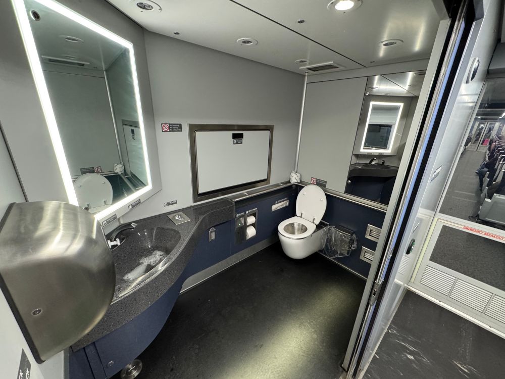A modern, compact restroom interior featuring a toilet, sink, and illuminated mirror. The walls are gray, and there are signs posted. The door to the restroom is partially open, showing a corridor with seating visible in the background.