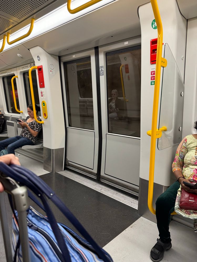 A view inside a subway car featuring two seated passengers. One woman is looking at her phone, while another sits across, holding her phone as well. The doors of the subway can be seen in the background, with yellow handrails and various informational signs