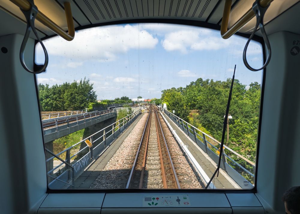 View from the front of a train, showing railway tracks extending into the distance with greenery on either side. A handrail is visible on the left side of the image, and the sky is partly cloudy.