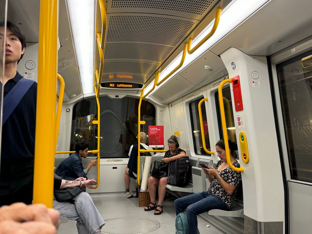 Interior of a subway train showing multiple passengers. The scene includes two women sitting on benches, one focused on her phone, and a man sitting nearby. The train has yellow handrails, and a display indicates the route to "Lufthaven."