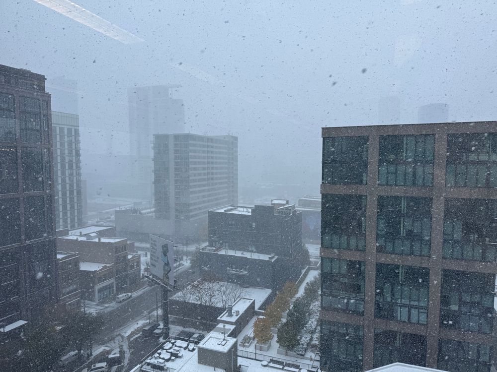 A snowy urban landscape with heavy snowfall obscuring visibility. Buildings and streets are covered in snow, creating a wintry scene.