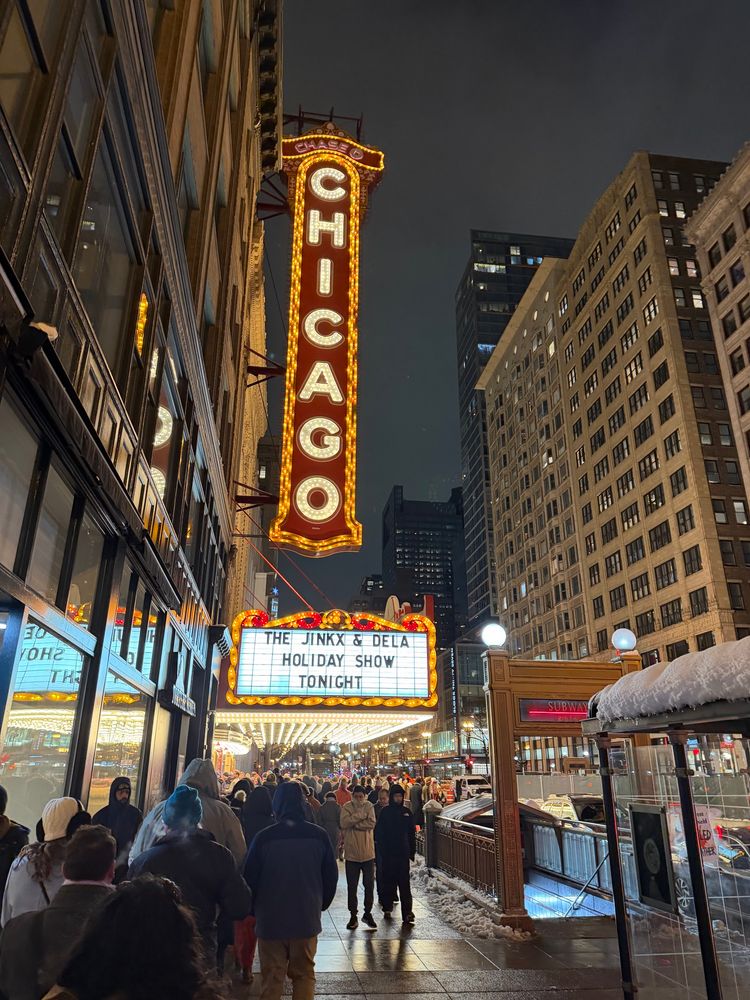The image features the Chicago Theater illuminated at night, showcasing its iconic marquee announcing "The Jinkx & Dela Holiday Show Tonight." A crowd of people is gathered outside, with snowy sidewalks and city buildings in the background.