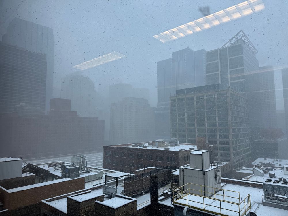 A snowy urban landscape seen through a window, with buildings fading into a white haze due to heavy snowfall. Rooftops are blanketed in snow, and visibility is low.