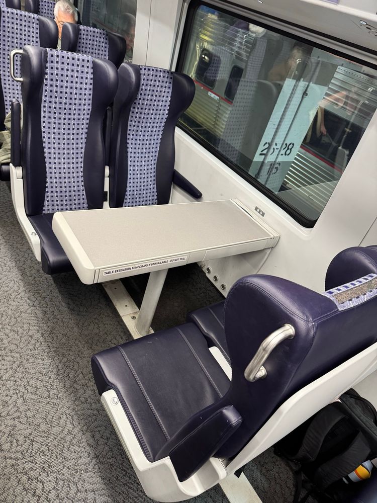 Interior of a train showing a set of four plush seating with a folding table between seats. A person is partially visible in the background. The decor features a modern design with a patterned backrest.