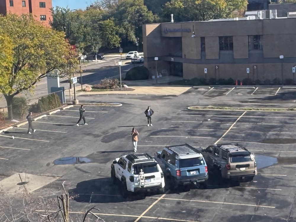 A parking lot with five vehicles parked, including three SUVs in a row. A group of four people is walking across the lot taking photos of the SUVs, with a few trees and a building in the background.
