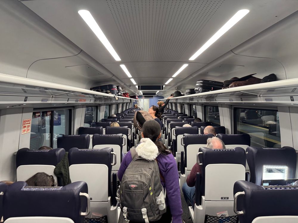 Interior view of a train car featuring rows of seats, with passengers standing and sitting. Luggage is stored overhead. The setting appears to be during travel, with visible windows showing a platform outside.